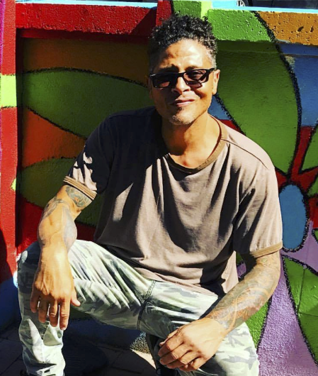 Photo of the artist in a t-shirt looking at the camera, kneeling on the ground and smiling behind sunglasses.