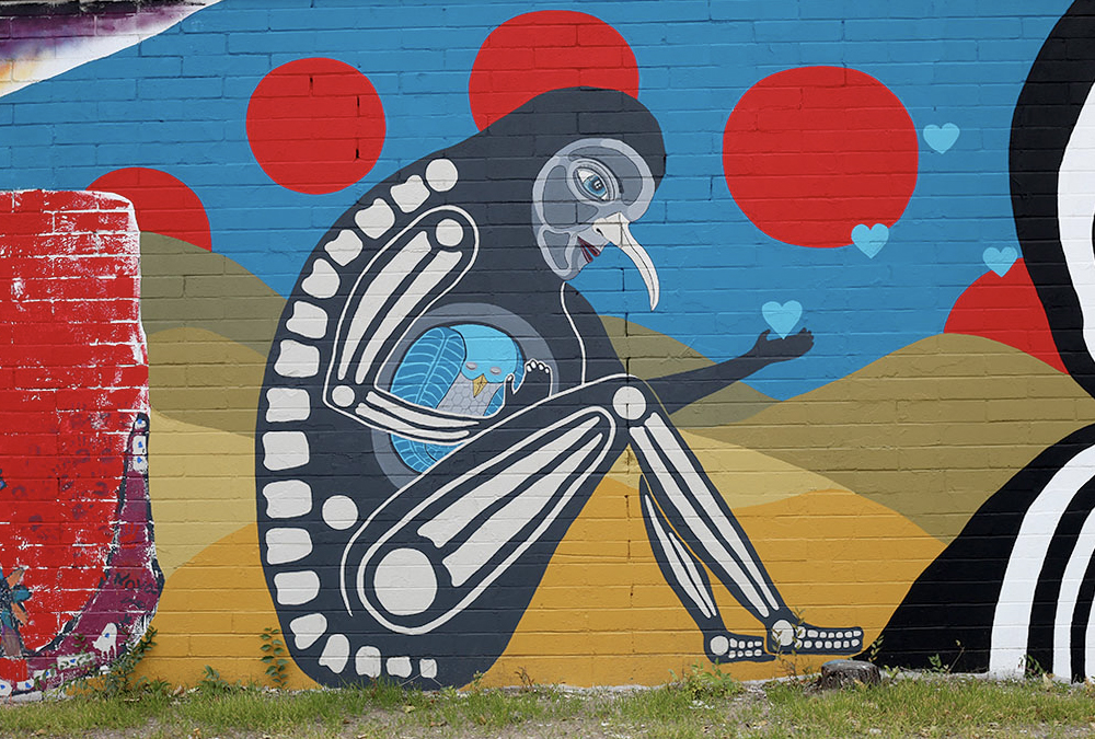 Photo of a mural of a strnge creature sitting down that is kind of a cross between a human sketeton body and a bird head with circles surrounding it.