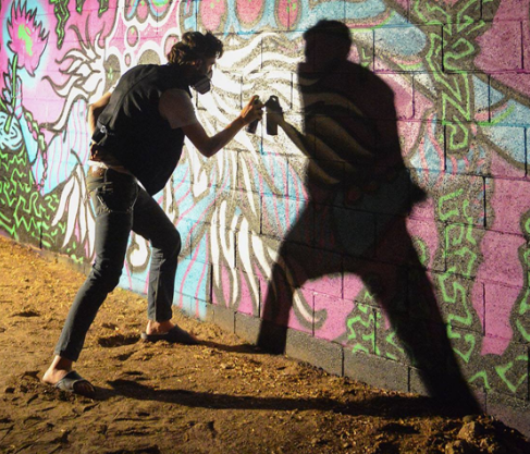 dramatic photo of the artist spray painting a wall in the dark of the night. his shadow is cast.