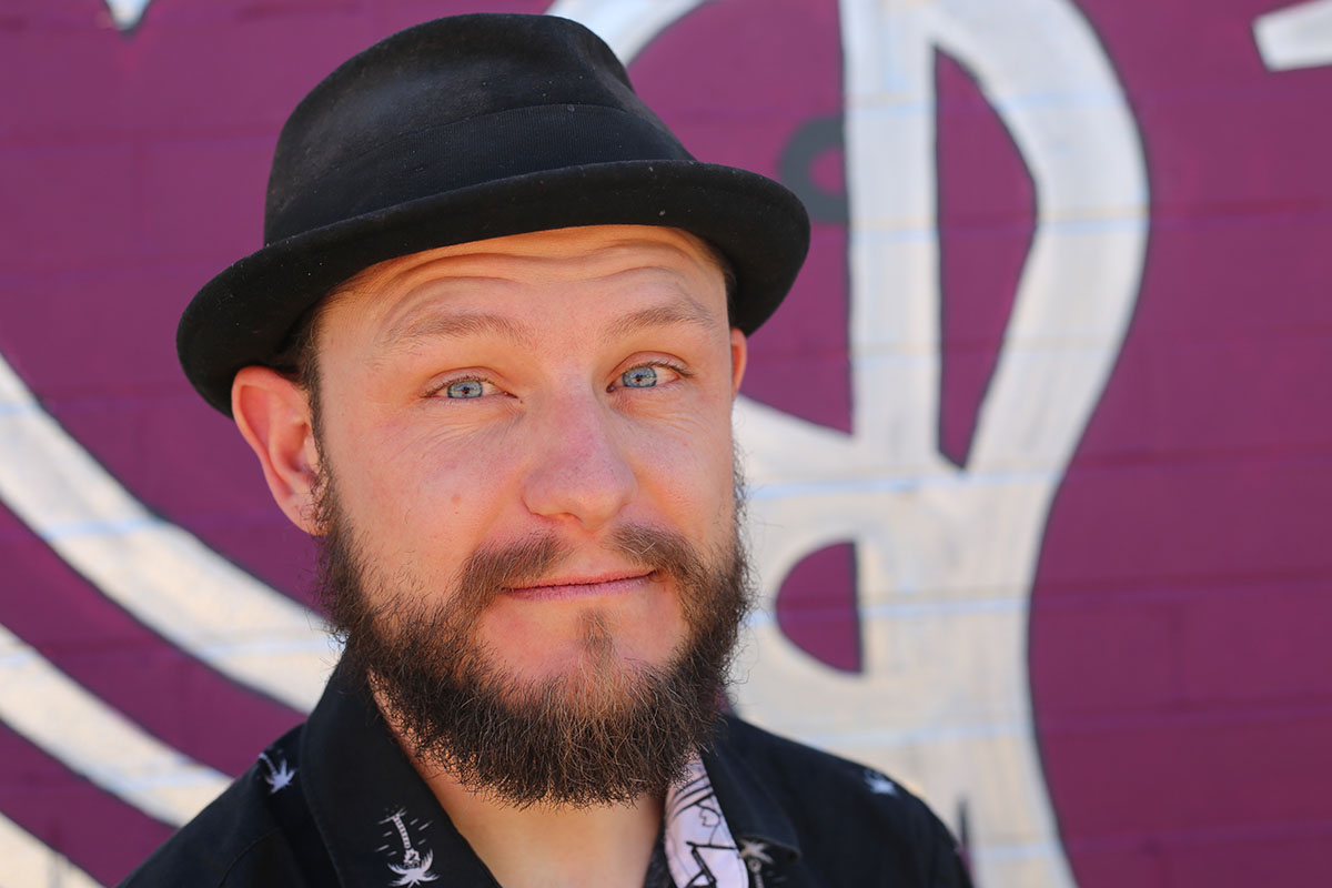 Photo of the blue eyed, bearded artist in a hat looking at camera smiling.